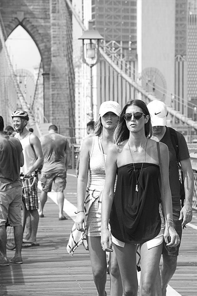 Black and white image of pedestrians walking on the upper-level of the Brooklyn Bridge, it&rsquo;s summer time, everyone in this image is wearing shorts